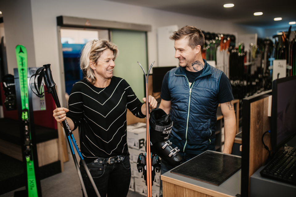 A woman is holding skis and poles in her hands, standing next to a man. Both are smiling.